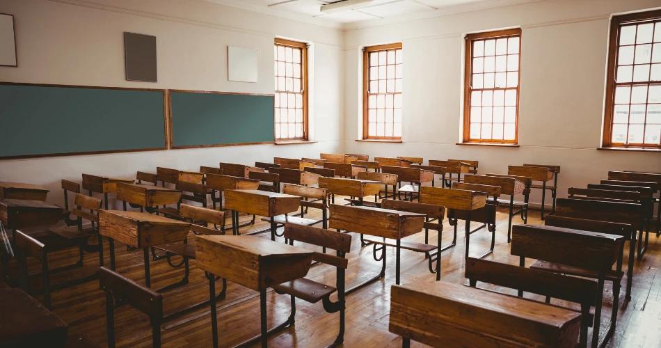 An empty classroom with wooden desks, green chalkboards, and sunlight streaming through large windows, symbolizing the impact of the NCTE recognition withdrawal on educational institutions.